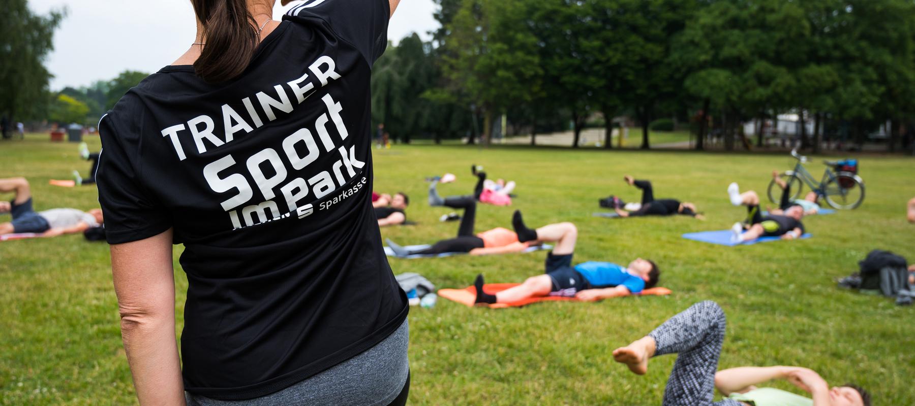 Trainerin mit schwarzem Shirt leitet Sportübungen im Park; Teilnehmer liegen auf Matten und trainieren im Hintergrund.